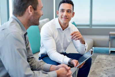 Two smiling male work coworkers sitting on a sofa in a modern office talking together over a digital tablet; Shutterstock ID 576773101; purchase_order: Brand Image Library Refresh Reconciliation Nov 2024