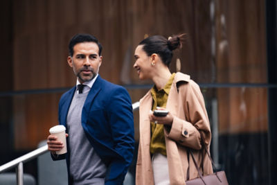 A stylish business couple walks along a city street. The man carries a coffee while the woman chats on her phone, both dressed in smart, modern attire and engaging in a casual business moment.